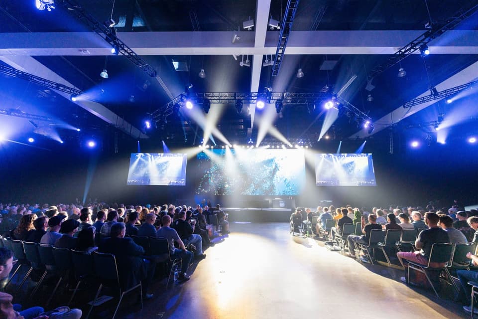 Large conference hall with audience facing a brightly lit stage and video screens